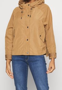 Brown hooded jacket with a layered texture, featuring snap buttons, side pockets, and a relaxed fit, paired with blue jeans.