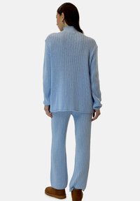 Jasnoniebieski sweter z prążkowanej dzianiny z wysokim kołnierzem, zestawiony z pasującymi spodniami z prążkowanej dzianiny. Modelka nosi brązowe kapcie. Miękka tekstura i luźny krój.