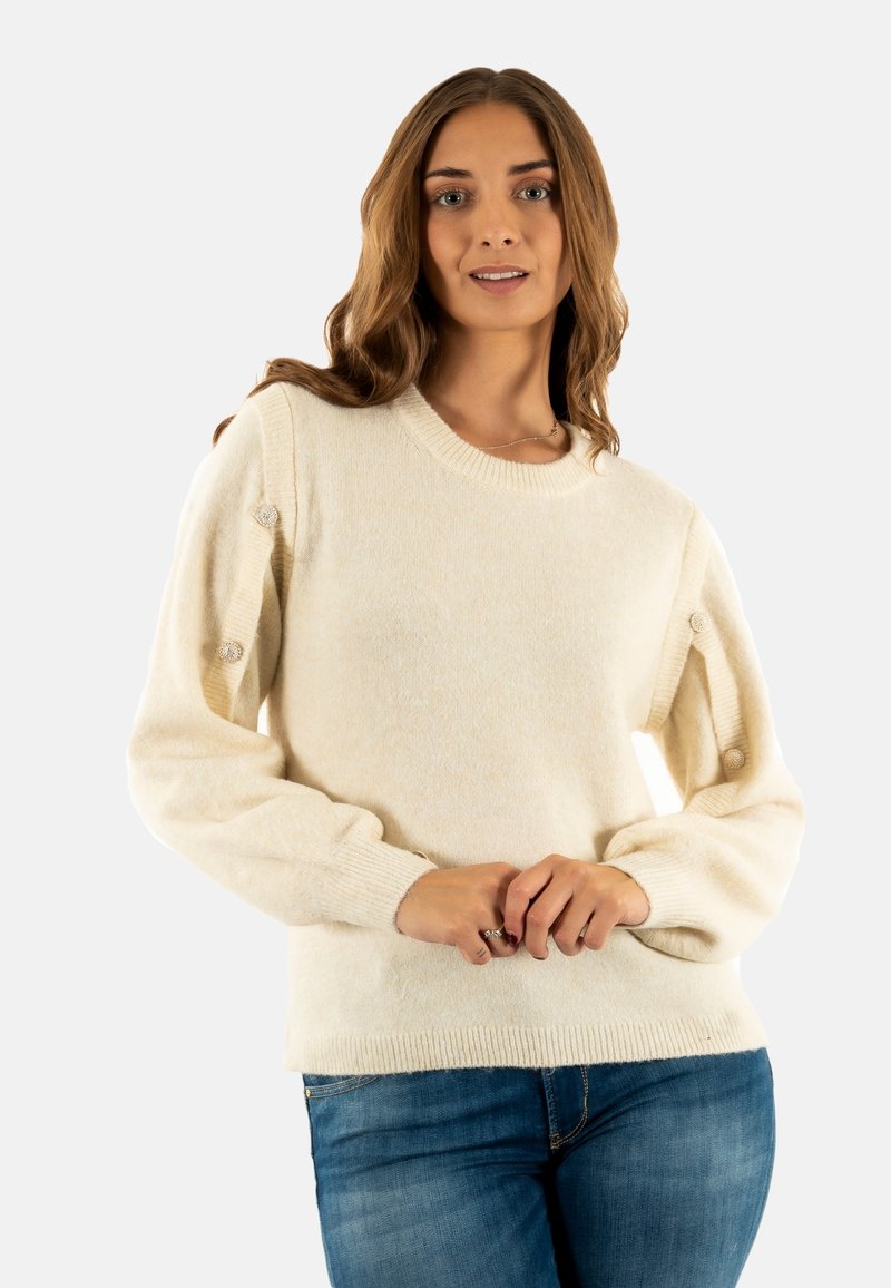 Jersey color crema con cuello redondo, tejido texturizado y botones decorativos en las mangas. Combinado con vaqueros de mezclilla.