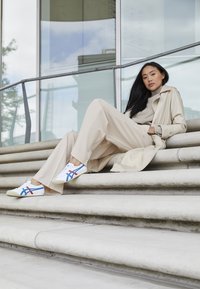 Beige trenchcoat, beige sweater, vide benede beige bukser, hvide sneakers med blå og røde accenter, siddende på stenmure ved siden af glasgelænder.