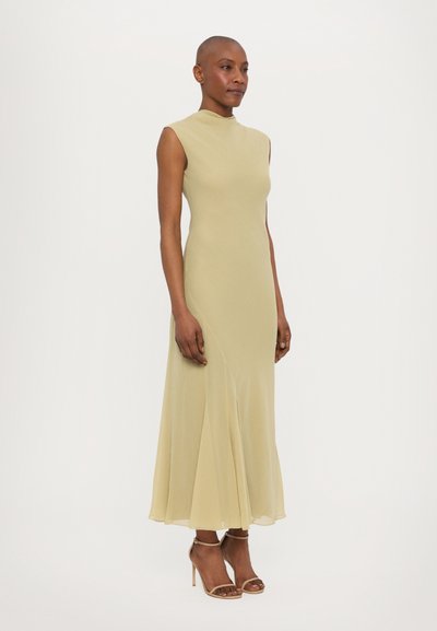 Mouwloze lichtgroene jurk met een hoge halslijn, getailleerd lijfje en een uitlopende rok, gecombineerd met beige sandaaltjes met banden, gladde stoftextuur.