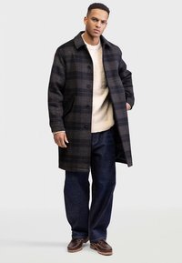 Schwarz-grau karierter Mantel mit einer strukturierten Silhouette. Mit Knopfleiste, Seitentaschen und weichem Innenfutter, kombiniert mit einem beigen Pullover.