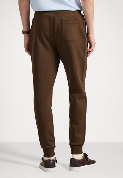 Los pantalones deportivos marrones cuentan con piernas ajustadas y un único bolsillo trasero. Los puños en los tobillos de canalé completan el diseño, combinados con zapatillas informales.