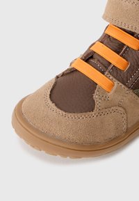 Brown and tan shoe with a suede exterior, leather accents, and orange elastic straps. Round toe with a textured rubber sole.