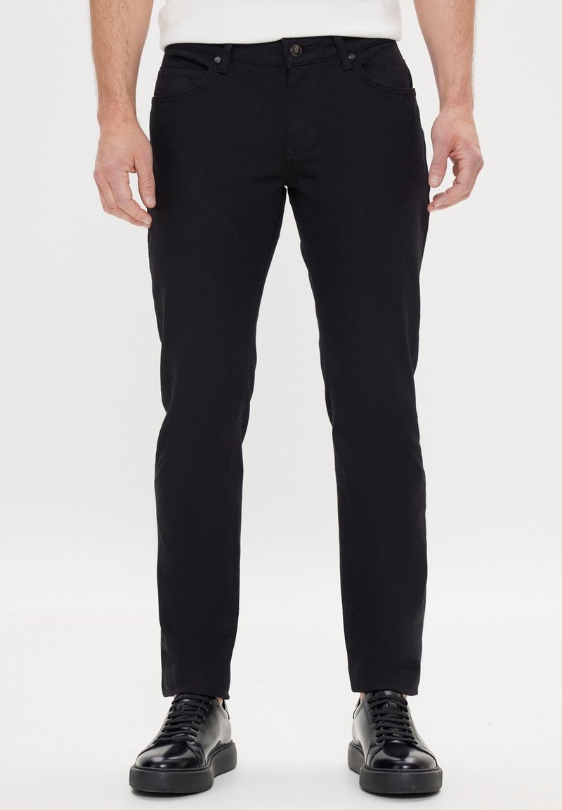 Černé džíny s úzkým střihem, vybavené předními kapsami, zapínáním na knoflíky a jemnými švy. Kombinovány s černými teniskami.