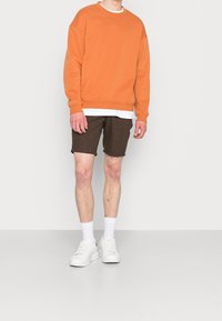 Sweatshirt orange avec col rond, sous-couche blanche, short marron, et baskets blanches, présentant une coupe décontractée et une texture visible.