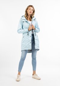 Lichte blauwe waterdichte jas met een elastische taille, rits- en drukknoopsluiting, en een capuchon; gecombineerd met licht denim jeans en beige schoenen.