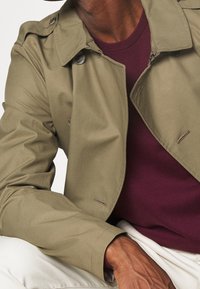 Olive green trench coat with a double-breasted design, featuring large buttons and a smooth texture, layered over a maroon sweater.