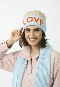 Gebreide beanie in lichtblauw en beige met "LOVE" in oranje. Draagt samen met een bijpassende lichtblauwe gestructureerde sjaal en een roze overhemden.