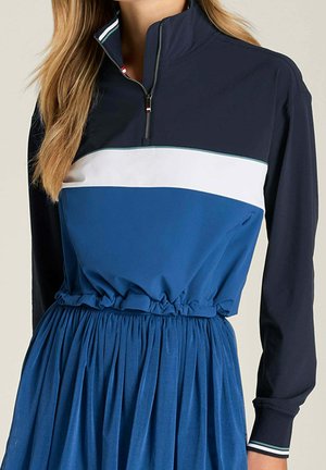 Donna che indossa un vestito a maniche lunghe blu, bianco e navy con collo con cerniera e vita arricciata.