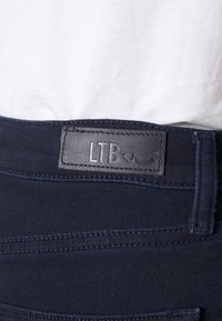 Mörkblå denim tyg med en mjuk textur. Har en svart läderetikett präglad med "LTB" på midjebandet. Framträdande sömmar synliga.