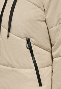 Doudoune beige avec fermeture éclair noire, arborant un motif matelassé diagonal, une texture de tissu contrastée et une capuche avec cordon de serrage.