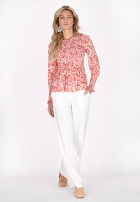 Top lungo a maniche lunghe di colore rosso con corpetto arricciato, abbinato a pantaloni bianchi a gamba larga e scarpe beige slip-on. Tessuto dalla consistenza liscia.