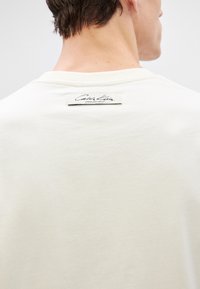 Beige Baumwoll-T-Shirt mit klassischem Rundhalsausschnitt und einem sichtbaren schwarzen Etikett auf der Rückseite, das "Calvin Klein, Designed in New York City" lautet.