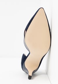 Chaussure à talon aiguille en daim bleu marine avec un nez pointu, présentant un talon texturé et une semelle lisse de couleur claire. Découpes latérales visibles.