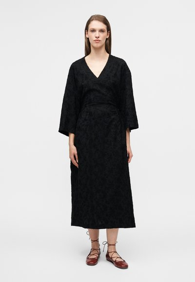 Robe portefeuille noire avec un col en V, des manches trois-quarts, un tissu texturé et un motif floral, associée à des chaussures plates marron à lacets.