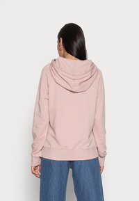 Felpa rosa con una vestibilità comoda e un ampio cappuccio, con polsini e orlo a coste. Indossata con pantaloni in denim blu a gamba larga.