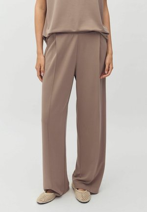 Personne portant un pantalon ample couleur taupe et un haut assorti, avec des chaussures beiges tissées sans lacets, debout devant un fond clair uni.