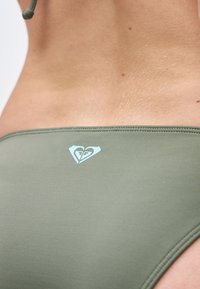 Κοντινό πλάνο του κάτω μέρους της πλάτης ενός ατόμου που φοράει μαγιό σε ελιά πράσινο χρώμα, με ένα μικρό λευκό λογότυπο σε σχήμα καρδιάς στο κέντρο της πίσω μέσης.