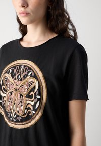 T-shirt nera con un design centrale di farfalla, caratterizzato da un ricamo intricato in oro, rosa e bianco. Maniche corte, tessuto morbido.