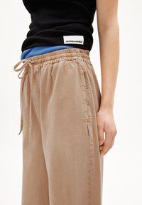 Pantalons de jogging beige en tissu doux, dotés d'une taille élastique avec cordon de serrage, de poches latérales et d'une petite étiquette de marque sur la couture latérale.