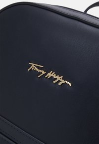 Tmavě modrá kožená taška s prošívanými okraji a zlatým kovovým logem "Tommy Hilfiger" na přední straně.