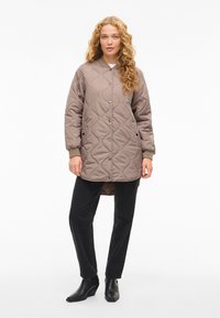 Pikowana, długorękawowa kurtka w kolorze taupe z zatrzaskami, bocznymi kieszeniami i ściągaczami na mankietach, zestawiona z czarnymi spodniami i botkami na obcasie.