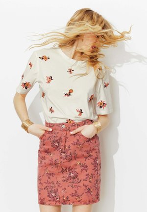 Femme aux cheveux blonds flottants portant un t-shirt blanc brodé de fleurs et une jupe taille haute rose à motifs floraux, les mains dans les poches.