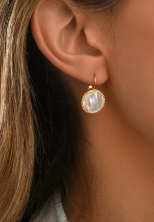 Gros plan d’une oreille portant une boucle d’oreille ronde en or avec un centre en nacre blanche et un bord doré orné de perles.