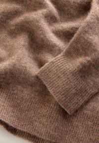 Brauner Strickpulloverstoff mit einer weichen, flauschigen Textur. Verfügt über gerippte Bündchen und einen lockeren Fall, der Variationen in der Farbtiefe zeigt.