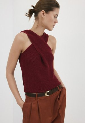 Kobieta ubrana w bezrękawnik w kolorze bordowym z krzyżującymi się ramiączkami i rdzawo-rdzawymi plisowanymi spodniami, ręce w kieszeniach, patrząca bokiem.