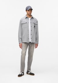 Camisa gris de botones con dos bolsillos en el pecho, superpuesta a una camiseta blanca. Se lleva con jeans gris claro y zapatos negros. Gorro gris en la cabeza.