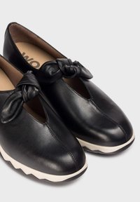 Czarne skórzane buty z okrągłym noskiem i elastycznym detalem w formie kokardy z przodu, z białą podeszwą w zygzakowaty wzór.