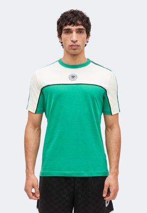 Nuori mies, jolla on lyhyet kiharat hiukset ja joka pukeutuu vihreään ja valkoiseen lyhythihaiseen paitaan, jossa on tunnus, seisoo yksivärisen vaalean taustan edessä.