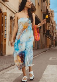 Sinine ja oranž abstraktse mustriga ilma olemasoleva rihmata kleit, valged jalatsid, roosa kootud kott, seismas tänaval, hoides kollast jääpulk. 