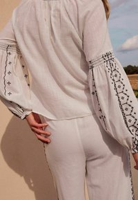 Hvid bluse med udblæste ærmer, der har sorte broderede mønstre langs manchetterne. Stoffet ser let og struktureret ud.