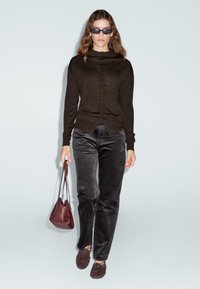 Donna che indossa un cardigan scuro con bottoni, pantaloni di velluto neri, occhiali da sole e mocassini marroni, mentre porta una piccola borsa rossa su uno sfondo semplice.