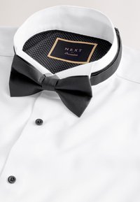 White dress shirt with black buttons and a black pre-tied bow tie under a white collar with a polka-dot inner lining and brand label.