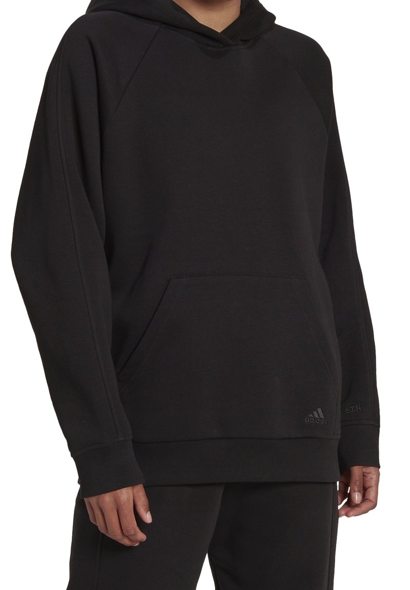 Personne portant un sweat à capuche Adidas noir uni avec poche frontale et poignets côtelés, montrée des épaules jusqu'au milieu des cuisses sur fond blanc.