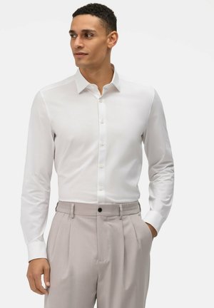 Camicia bianca a maniche lunghe con colletto a punta, bottoni frontali e texture liscia, abbinata a pantaloni grigi chiari a pieghe.
