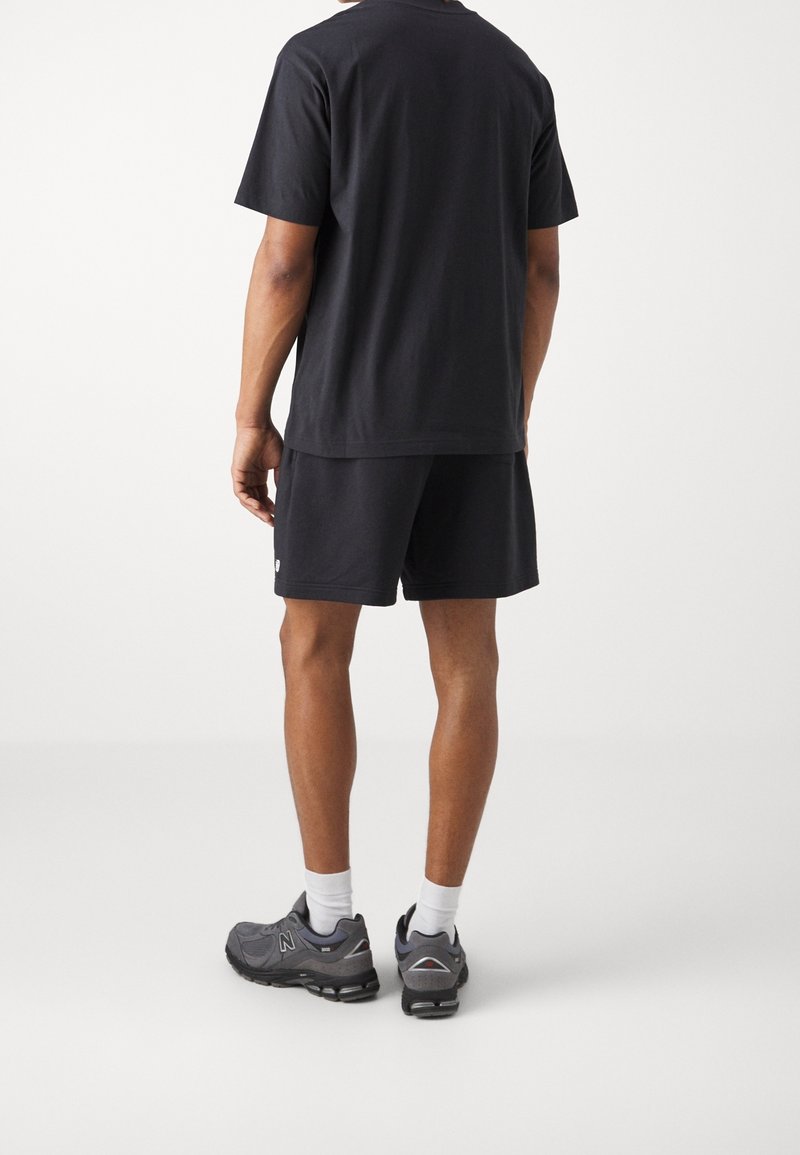 Homme debout, dos tourné, portant un t-shirt noir à manches courtes, un short noir, des chaussettes blanches et des chaussures de course grises sur fond blanc uni.