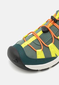 Grønn og grå treningssko med mesh-materiale, lyse gule og oransje snørebånd, gummitåhette og teksturerte paneler for grep.
