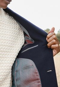 Abrigo de lana azul marino con interior de forro estampado; cuenta con bolsillos y acentos grises. Se lleva con un suéter texturizado color crema.