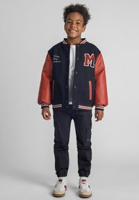 Schwarze und rote College-Jacke mit Druckknöpfen, ausgestattet mit einem gestickten "M" und weißen Buchstaben, kombiniert mit dunklen Cargo-Hosen und Sneakers.