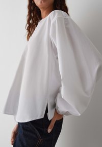 Weiße Bluse mit langen Ärmeln, runder Ausschnitt, gerafften Ärmeln, seitlichen Schlitzen und glatter Textur, kombiniert mit dunklen Jeans.