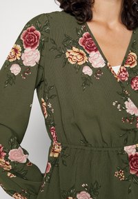 Zielona sukienka w fasonie kopertowym z kwiatowym nadrukiem, przedstawiająca duże róże i piwonie w odcieniach różu, żółci i burgundu. Teksturowany materiał i elastyczna talia.