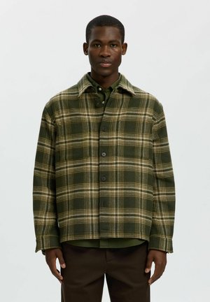 Man wears green and beige plaid buttoned jacket over a green shirt and dark brown pants against a plain light background.
