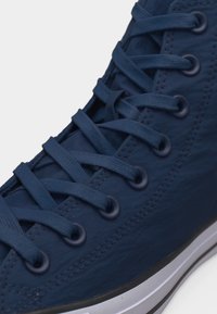 Αθλητικό παπούτσι μπλοκ υψηλού τύπου σε ναυτικό μπλε με αντιστοιχισμένα κορδόνια περασμένα μέσα από μεταλλικά ρουχιστικά, υφασμάτινο με υφή και λευκή ελαστική σόλα με μαύρο περίγραμμα.