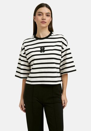 Woman wearing a black and white horizontal striped short-sleeve shirt with a black logo on the chest, paired with high-waisted black pants.