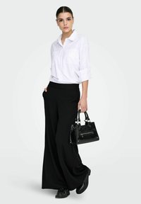 Bílá košile na knoflíky s vyhrnutými rukávy, černé široké kalhoty s proužky a černá texturovaná kabelka. Outfit doplňují černé boty.